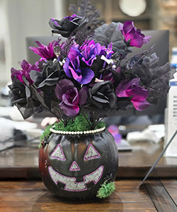 Pumpkin entry #7 - black pumpkin vase with white eyes, nose, and mouth which are outlined in purpler. Pearls line to cut out edge at the top of the pumpkin and purple/black flowers coming out of the top
